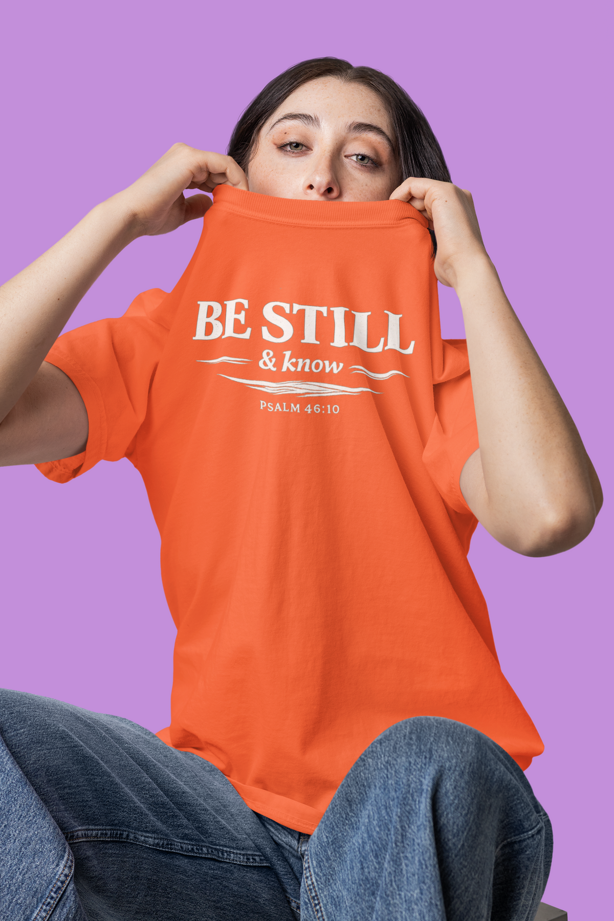 Person wearing an orange t-shirt with 'BE STILL & KNOW' text on a purple background