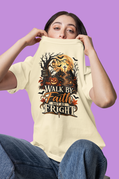 A woman holding up an ivory-colored t-shirt with a graphic design that reads 'Walk by Faith Not by Fright' in a spooky Halloween theme, set against a solid purple background.