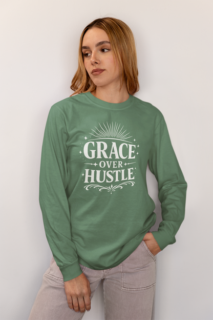 A woman wearing a sage green long-sleeve crewneck sweatshirt with a white graphic print that reads 'GRACE OVER HUSTLE' on the front, paired with light gray jeans, standing against a plain light gray wall.