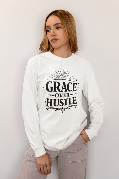 A woman wearing a white long-sleeve crewneck sweatshirt with a black graphic print that reads 'GRACE OVER HUSTLE' with decorative sunburst and scroll details, paired with light gray pants, set against a plain light gray wall.