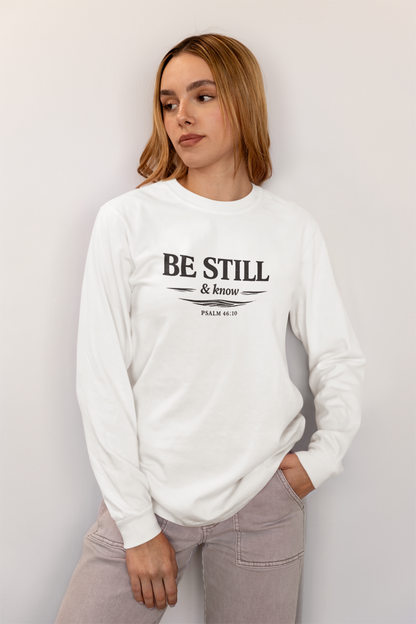 A woman wearing a white long-sleeve crewneck sweatshirt with the black printed text 'BE STILL & KNOW' and 'PSALM 37:4' on the front, paired with light gray jeans, posing against a light gray wall.