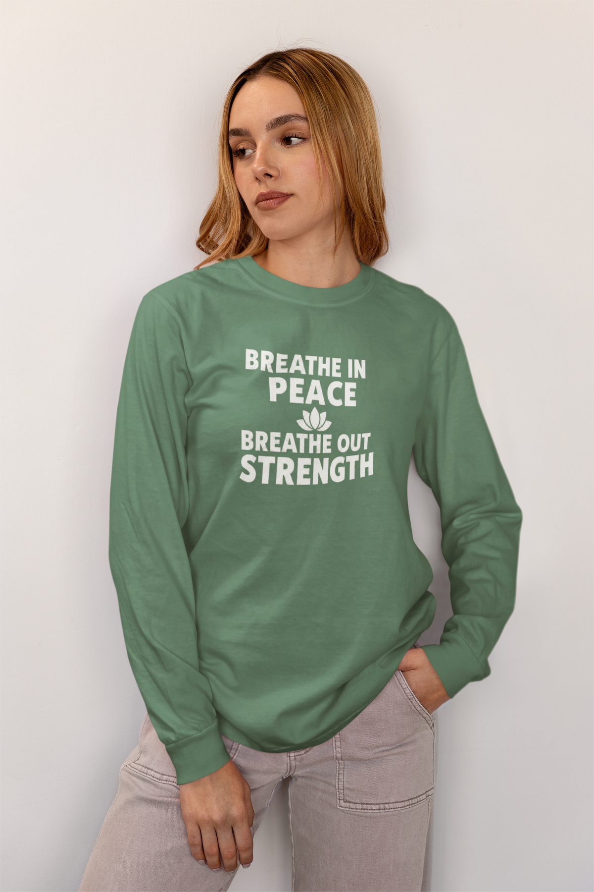 A woman wearing a sage green long-sleeve t-shirt with the white printed text 'BREATHE IN PEACE BREATHE OUT STRENGTH' and a small lotus flower graphic, paired with light gray jeans, standing against a plain light gray wall.