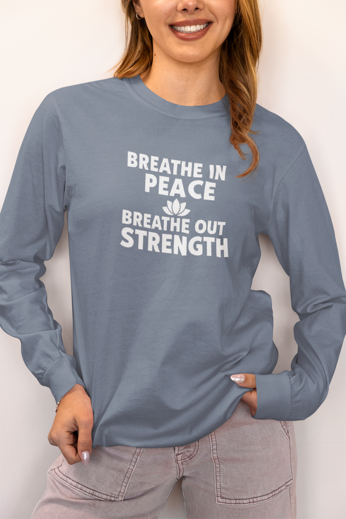 A woman wearing a slate blue long-sleeve t-shirt with a white graphic print that reads 'BREATHE IN PEACE BREATHE OUT STRENGTH' with a small lotus flower icon, paired with light gray jeans.