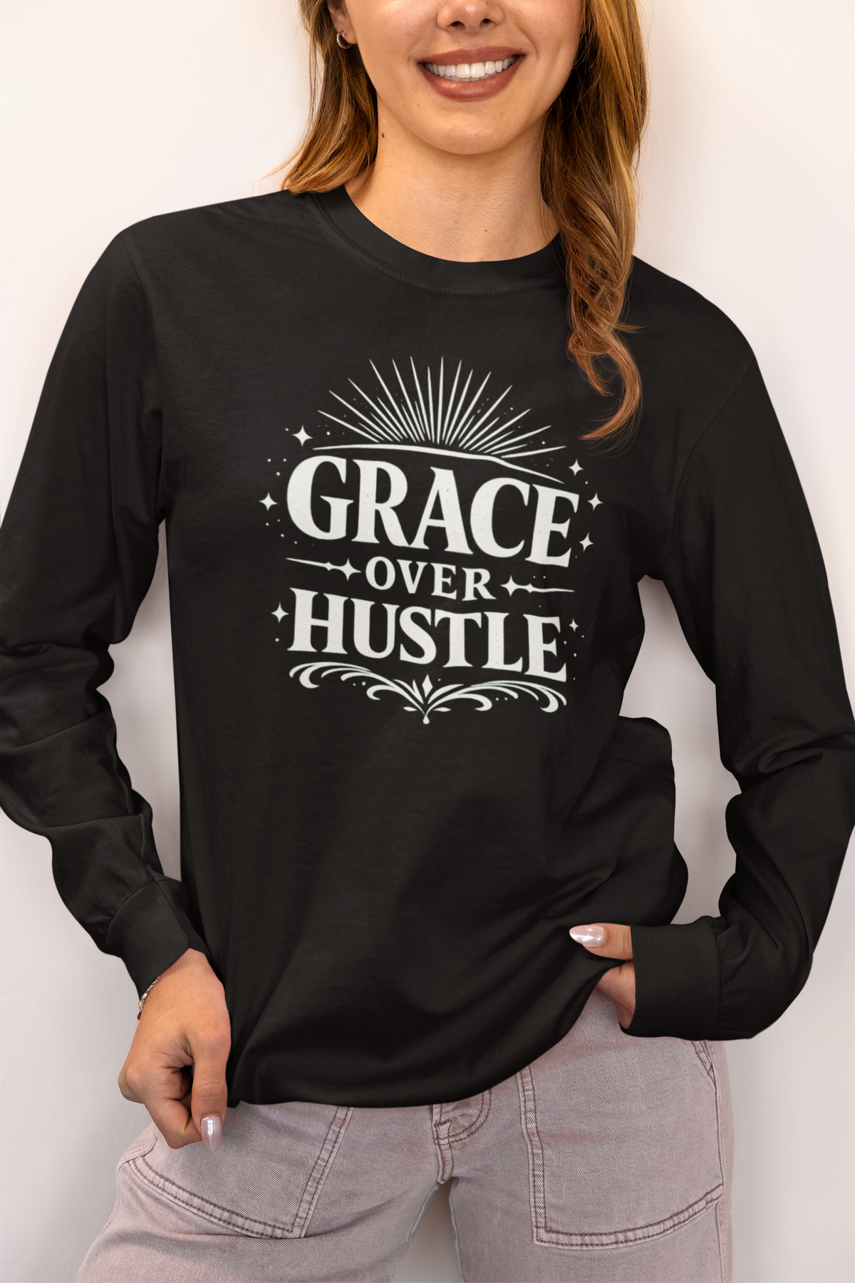 A woman wearing a black long-sleeve t-shirt with a white graphic print that reads 'GRACE OVER HUSTLE' in a bold typewriter font with decorative sunburst and scroll details.