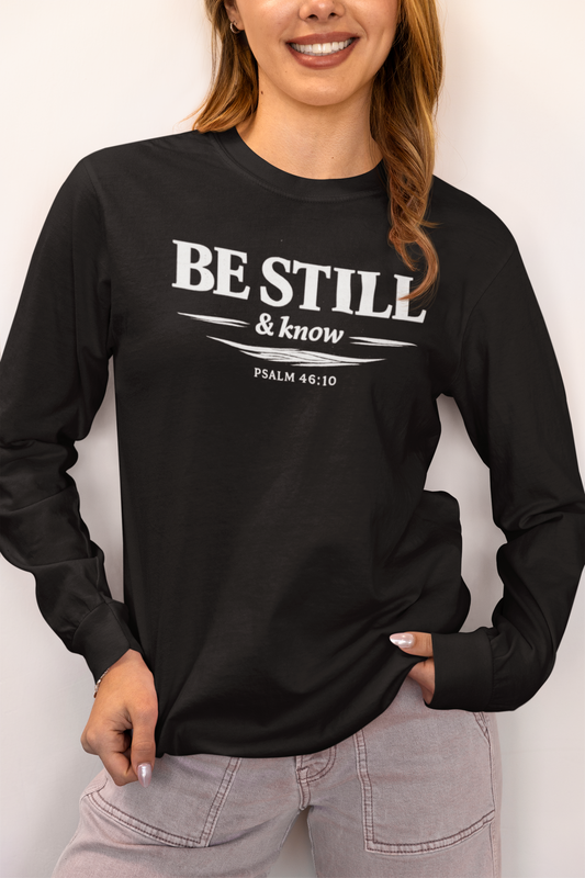 A woman wearing a black long-sleeve t-shirt with a white graphic print that reads 'BE STILL & know PSALM 46:10' on the front, paired with light gray jeans, against a plain off-white background.
