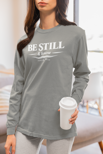 A woman wearing a gray long-sleeve t-shirt with the text 'BE STILL & know PRIMAL AID' printed in white on the front, holding a white coffee cup in a brightly lit room.