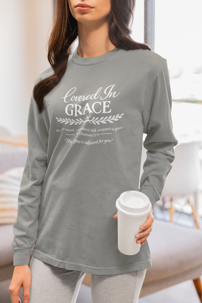 A woman wearing a heather gray long-sleeve graphic t-shirt with 'Covered In Grace' printed in white on the front, holding a white coffee cup in a brightly lit room.