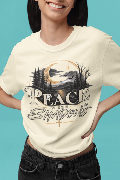 A woman wearing an ivory-colored graphic t-shirt with a forest and moon design that reads 'PEACE IS THE SHADOWS', paired with black jeans, set against a solid teal background.