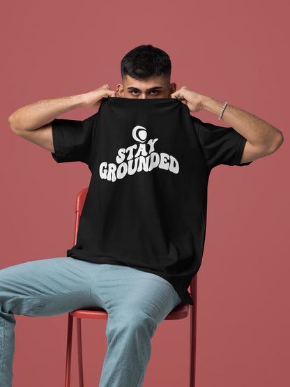 Person wearing a black t-shirt with 'Stay Grounded' text, sitting on a red chair against a pink background