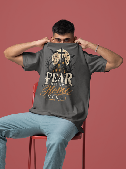 A man sitting on a red chair holds up a pepper t-shirt with a Halloween graphic that reads 'FEAR HAS NO HOME HERE' in white and gold text, set against a solid dusty rose background.
