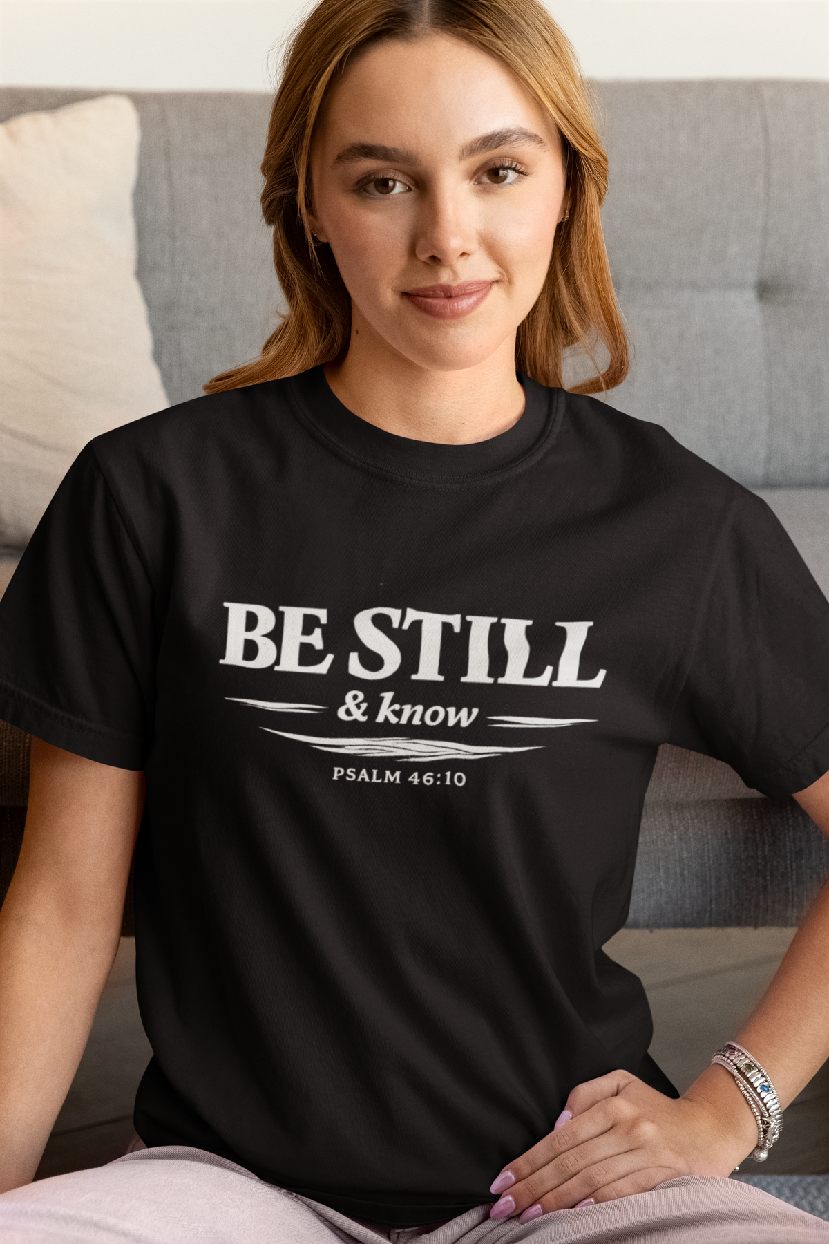 Woman wearing a black t-shirt with 'Be Still & Know' text, sitting on a couch.