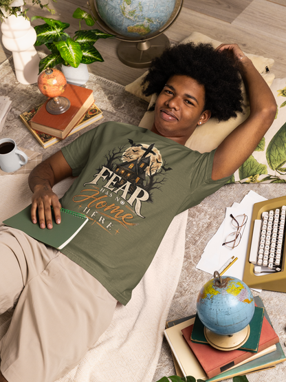 A man lies on a beige blanket in a cozy, book-filled room, wearing an moss graphic t-shirt with the text 'FEAR HAS NO HOME HERE' and a graphic of a house and cross. He is surrounded by books, globes, plants, and a coffee mug.