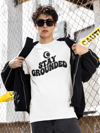 Person wearing a white t-shirt with 'Stay Grounded' text and a black jacket, standing against a chain-link fence.