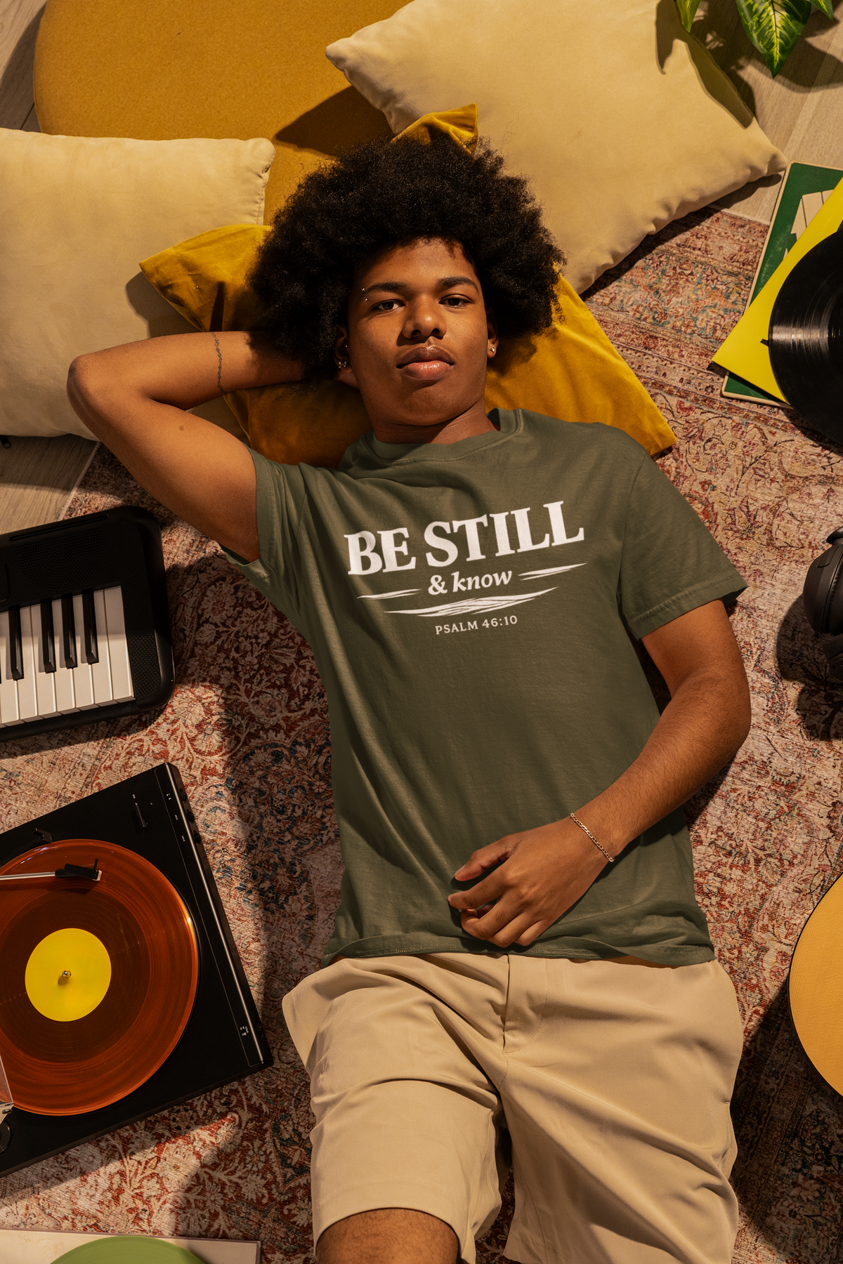 Person wearing a 'Be Still & Know' shirt lying on a patterned rug with musical instruments around