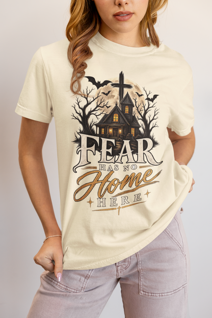 A woman wearing an ivory graphic t-shirt with a spooky haunted house design and the text 'FEAR HAS NO HOME HERE', paired with light gray jeans, against a neutral studio background.