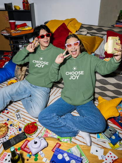Two people wearing light green 'CHOOSE JOY' hoodies sitting on a couch with a game board and snacks around.