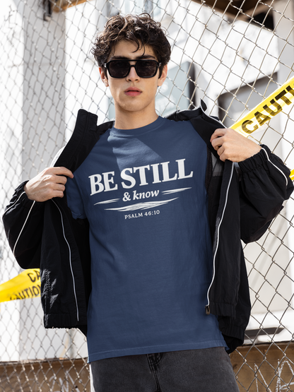 Person wearing a blue 'BE STILL & KNOW' t-shirt with a black jacket in front of a chain-link fence.