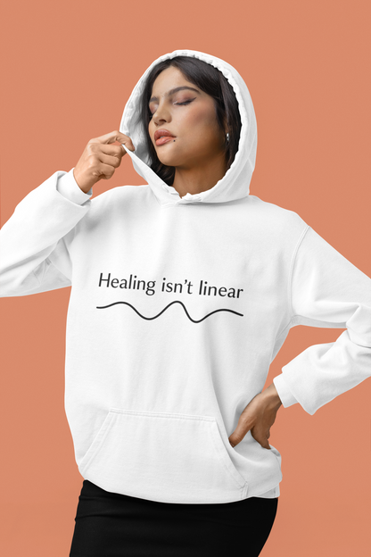 Person wearing a white hoodie with 'Healing isn't linear' text on a peach background