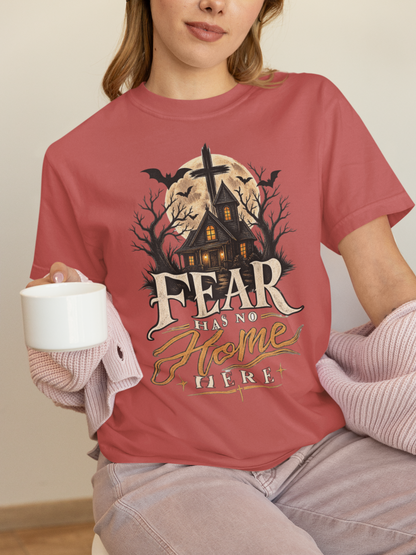 A woman wearing a crimson graphic t-shirt with a Halloween haunted house design and the text 'FEAR HAS NO HOME HERE'. She is holding a white mug and wearing a pink knit sweater draped off her shoulders, paired with light gray jeans.