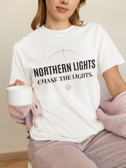 Person wearing a white 'Northern Lights – Chase the Lights' garment-dyed tee, holding a mug on a couch.