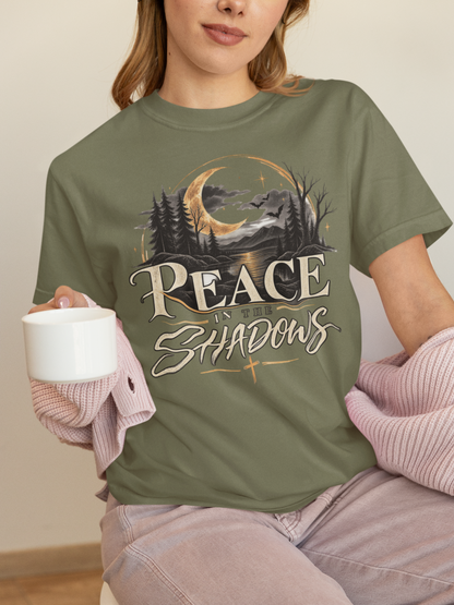 A woman wearing an olive green (moss) graphic t-shirt with a 'Peace in the Shadows' forest and moon design, holding a white mug and wearing a pink knit sweater draped off her shoulders.
