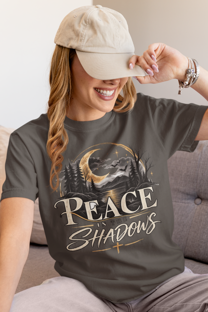A woman wearing a pepper graphic t-shirt with a 'PEACE IN THE SHADOWS' mountain and moon design, paired with a beige baseball cap, sitting on a gray couch.