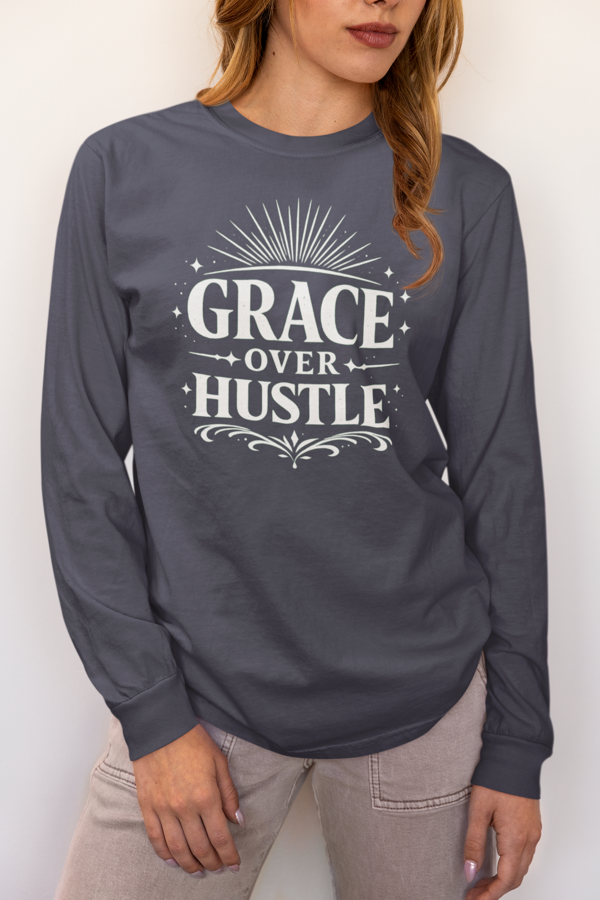 A woman wearing a dark slate gray long-sleeve t-shirt with a white graphic print that reads 'GRACE OVER HUSTLE' with decorative sunburst and scroll details, paired with light beige pants.