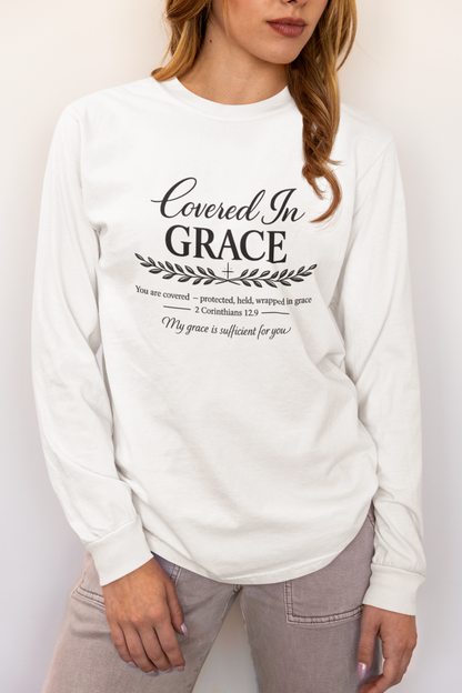 A woman wearing a white long-sleeve graphic t-shirt with black text that reads 'Covered In GRACE' and scripture text below, paired with light gray jeans.