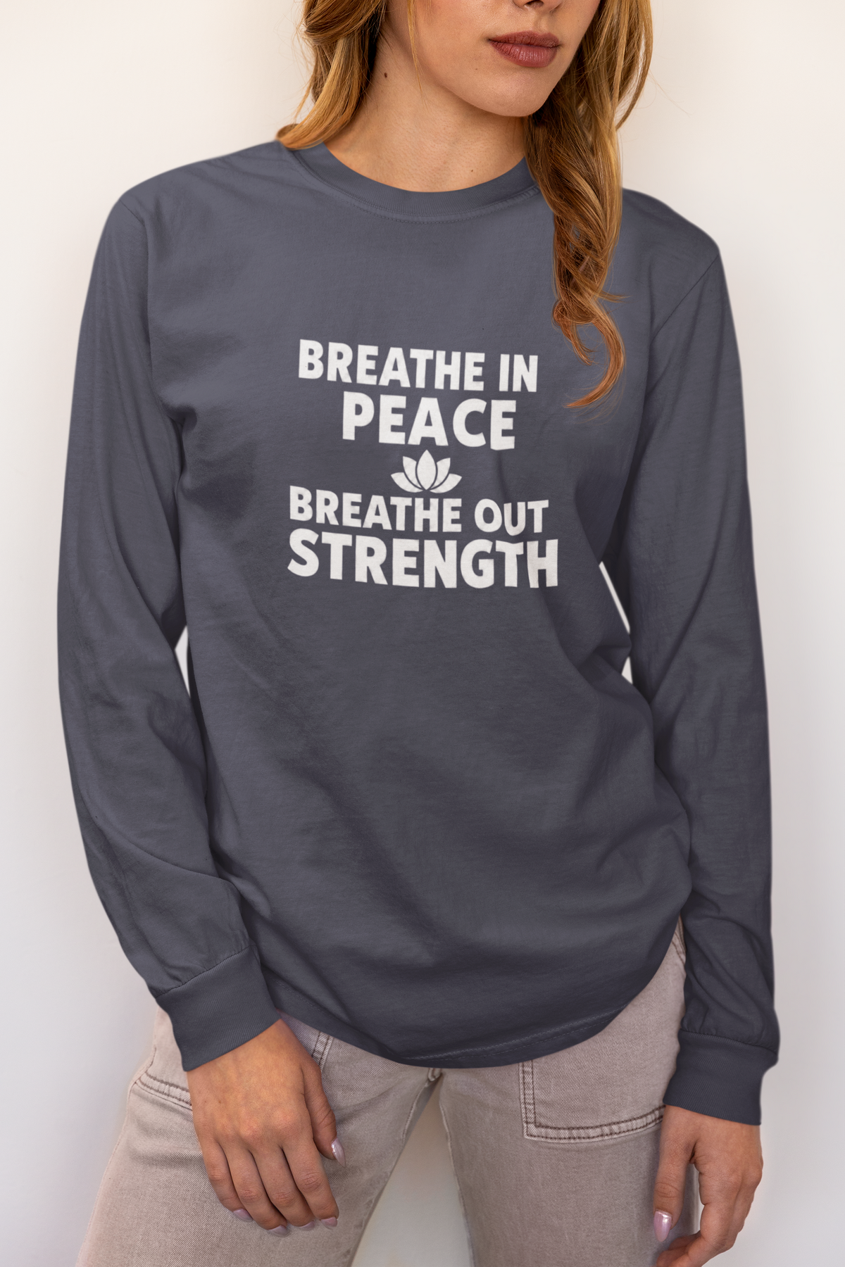 A woman wearing a dark gray long-sleeve t-shirt with the white printed text 'BREATHE IN PEACE BREATHE OUT STRENGTH' and a small lotus flower graphic on the front, paired with light gray jeans.