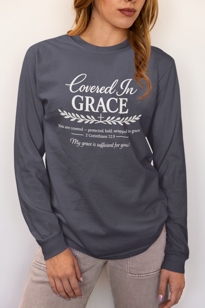 A woman wearing a dark gray long-sleeve t-shirt with a white graphic print that reads 'Covered In GRACE' along with scripture text, paired with light gray jeans, against a plain off-white background.