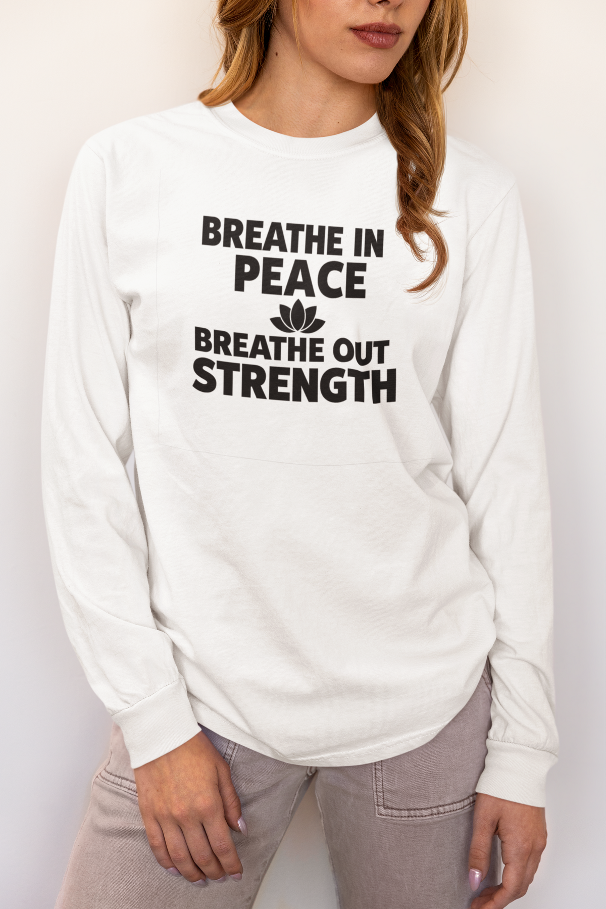 A woman wearing a white long-sleeve crewneck t-shirt with the black printed text 'BREATHE IN PEACE BREATHE OUT STRENGTH' and a small lotus flower graphic on the front, paired with light gray jeans.