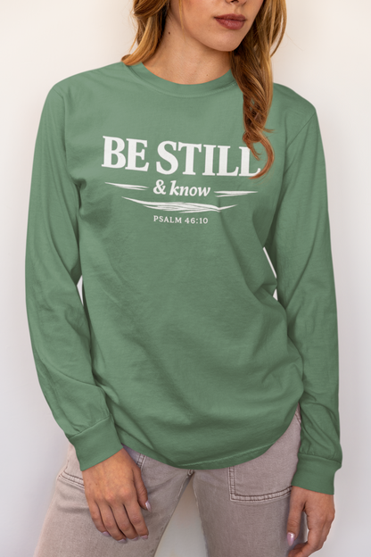A woman wearing a sage green long-sleeve t-shirt with a white graphic print that reads 'BE STILL & know PSALM 46:10'.