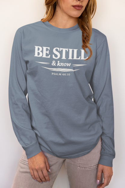 A woman wearing a dusty slate blue long-sleeve t-shirt with a white graphic print that reads 'BE STILL & know PSALM 46:10'.