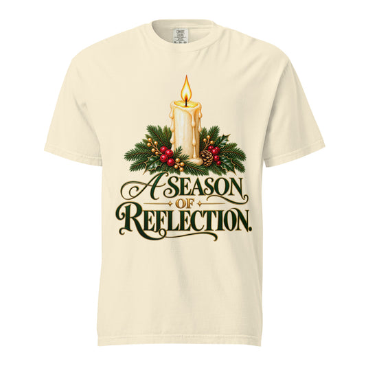 Front view of an Ivory “A Season of Reflection” Christmas tee featuring a lit candle, evergreen branches, red berries, a pinecone, and green-and-gold lettering.