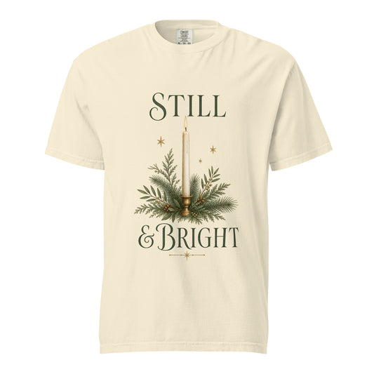 Front view of an Ivory Still & Bright Christmas tee featuring a taper candle, evergreen sprigs, gold stars, and forest-green lettering.