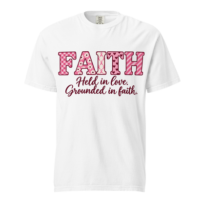White T-shirt with pink patterned FAITH lettering, awareness ribbon motifs, and burgundy “Held in love. Grounded in faith.” script.